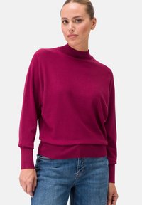 Fuchsia Strickpullover mit hohem Kragen und langen, weiten Ärmeln. Mit geripptem Saum und Bündchen, kombiniert mit blauen Jeans. Glatte Textur.