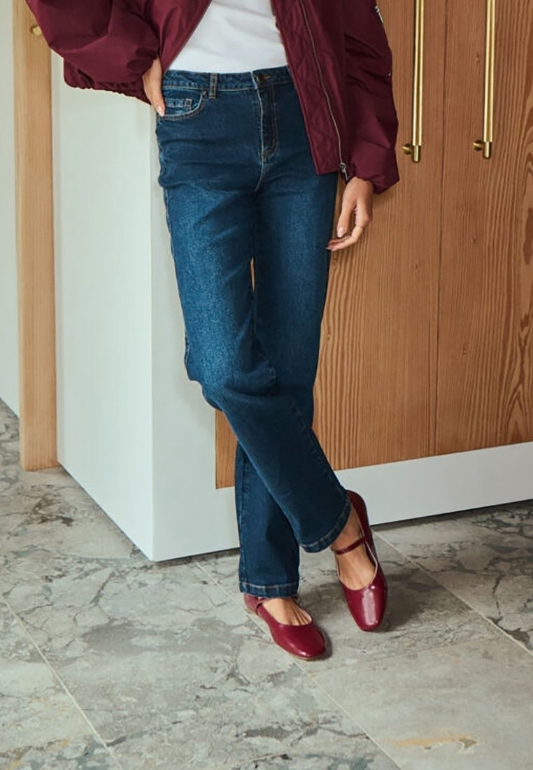 Femme appuyée contre le plan de travail de la cuisine portant un jean bleu foncé, une veste bordeaux, un haut blanc et des chaussures Mary Jane bordeaux sur un sol carrelé.