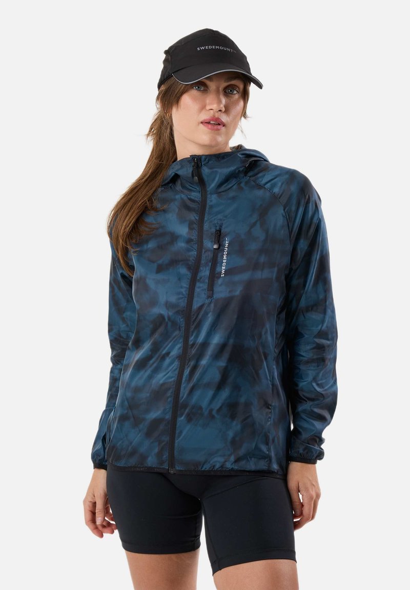 Femme portant une casquette noire et une veste zippée bleue à motif camouflage avec capuche, associée à un short noir ajusté, debout devant un fond blanc.