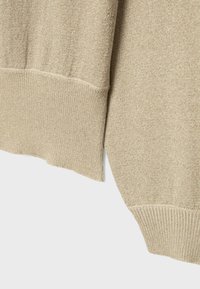 Posnetek v bližini bež pletenega puloverja z rebrastimi manšetami in subtilnim kovinskim nitkanjem na enobarvnem ozadju.