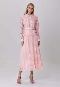 Femme portant une veste en dentelle rose clair avec des boutons en perles et une jupe midi plissée assortie, accompagnée de talons à bride de cheville blancs.