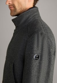 Anthrazitgrauer Wollmantel mit hohem Kragen, Fischgrätmuster und einem runden, schwarzen Logo auf dem linken Ärmel. Verfügt über frontale Taschen.