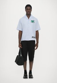 Camicia a maniche corte, a righe blu chiaro con testo e grafiche, abbinata a pantaloni cargo neri e una borsa nera; il modello è in posizione eretta.
