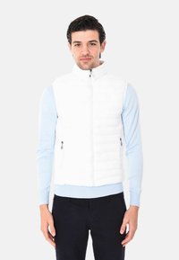 Gilet bianco imbottito con collo alto e chiusura lampo frontale. Due tasche laterali con chiusura a zip. Abbinato a una camicia a maniche lunghe azzurra.
