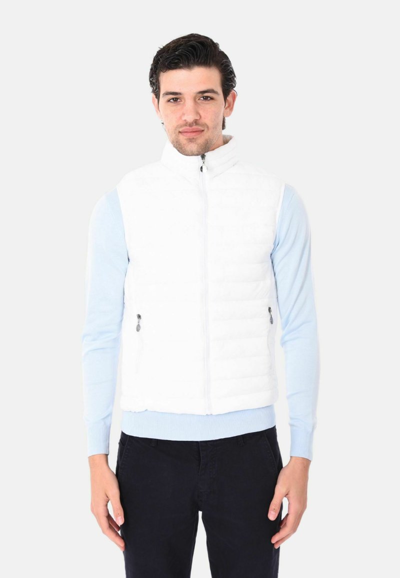 Gilet bianco imbottito con collo alto e chiusura lampo frontale. Due tasche laterali con chiusura a zip. Abbinato a una camicia a maniche lunghe azzurra.