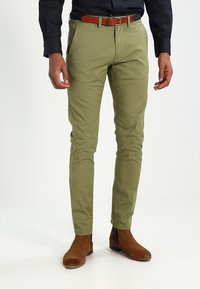 Green cotton chinos with a slim fit, brown leather belt, and brown suede ankle boots. Smooth texture and clean lines throughout.