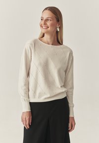 Világos bézs pulóver gyémántmintával és apró díszítésekkel, széles nyakkivágással és bordás mandzsettával, fekete alsórészekkel párosítva.