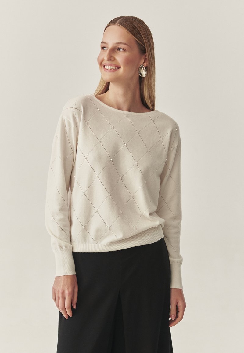 Világos bézs pulóver gyémántmintával és apró díszítésekkel, széles nyakkivágással és bordás mandzsettával, fekete alsórészekkel párosítva.