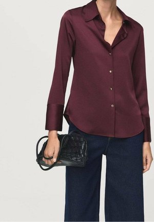 Kobieta w bordowej bluzce na guziki z długim rękawem i ciemnych, szerokich spodniach, trzymająca małą czarną plecioną skórzaną torebkę.