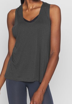 Detail osoby nosící tmavě šedé volné tílko bez rukávů a tmavě modré legíny na jednobarevném pozadí.