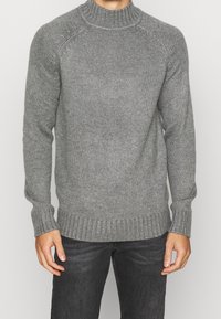 Maglione grigio a maglia con collo alto a costine, polsini a costine e finitura strutturata. Indossati jeans scuri.