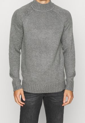 Grauer Strickpullover mit hochribbeligem Kragen, gerippten Bündchen und strukturiertem Finish. Dunkle Jeans getragen.