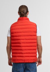 Rote Steppweste mit horizontalen Nähten, hohem Kragen und ohne Ärmel. Hat eine glatte Textur und wird über einem weißen T-Shirt getragen.