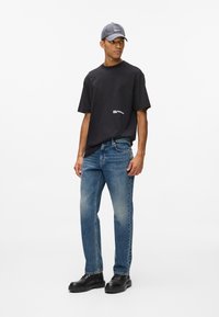 Juodas medvilnės marškinėliai su logotipu, šviesiai mėlyni džinsai ir juodi batai. Modelis dėvi pilką kepurę su kvadratiniu pleistru.