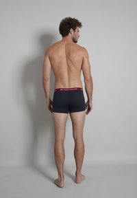 Boxers en jersey marine foncé, en matériau doux, avec une ceinture élastique rouge arborant un logo. Le modèle se tient pieds nus, vu de dos.