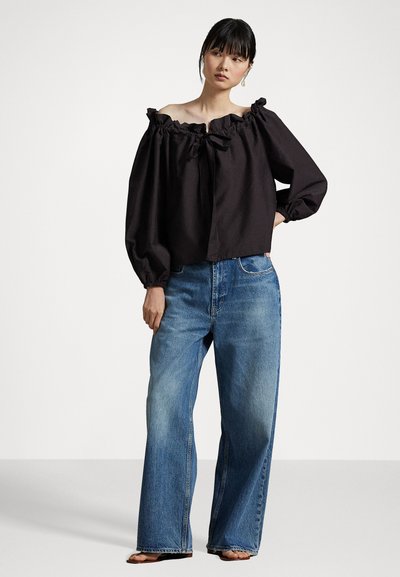Black off-the-shoulder blouse with ruffled neckline, tied bow detail, and long puff sleeves, paired with wide-leg light blue jeans.