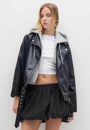 Jeune femme aux cheveux blonds portant une veste en cuir noir superposée sur un sweat à capuche gris, un crop top blanc et une jupe noire.
