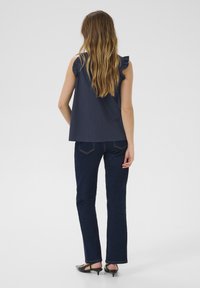 Top sans manches marine avec des détails à volants sur les épaules, associé à un pantalon en denim foncé évasé et des talons noirs, vu de dos.