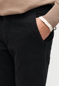 Μαύρο βαμβακερό παντελόνι με πλαϊνές τσέπες, λεία υφή, εφαρμοστή γραμμή, λεπτομέρεια μανσετών σε ταμπά και λευκό στο μανίκι, χωρίς εμφανή λογότυπα ή διακοσμητικά στοιχεία.