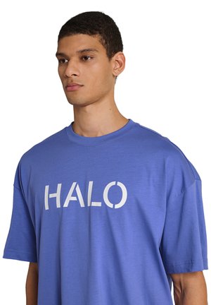 Junger Mann mit kurzem Haar, der ein locker sitzendes blaues T-Shirt mit dem weißen Aufdruck "HALO" trägt und leicht zur Seite schaut.