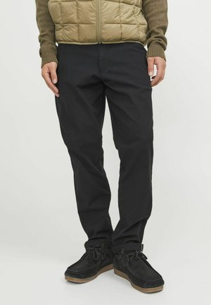 Schwarze Baumwollhose mit geradem Schnitt, kombiniert mit einer hellbraunen, gepolsterten Weste. Das Modell trägt schwarze Wildlederschuhe mit Gummisohle.