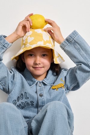 Sárga virágos bucket kalap, farmerdzseki "COOL KIDS" felirattal és tigris hímzéssel, citromot tartva, egy fehér fal előtt ülve.