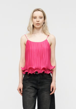 Donna che indossa una maglia smanicata plissettata di un rosa acceso con orlo arricciato e jeans neri a vita alta, in piedi contro uno sfondo chiaro uniforme.