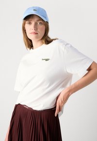 Hvit kortermet t-skjorte med grønt Lacoste-logo, kombinert med et brun plissert skjørt. Lyseblå caps på modellens hode.