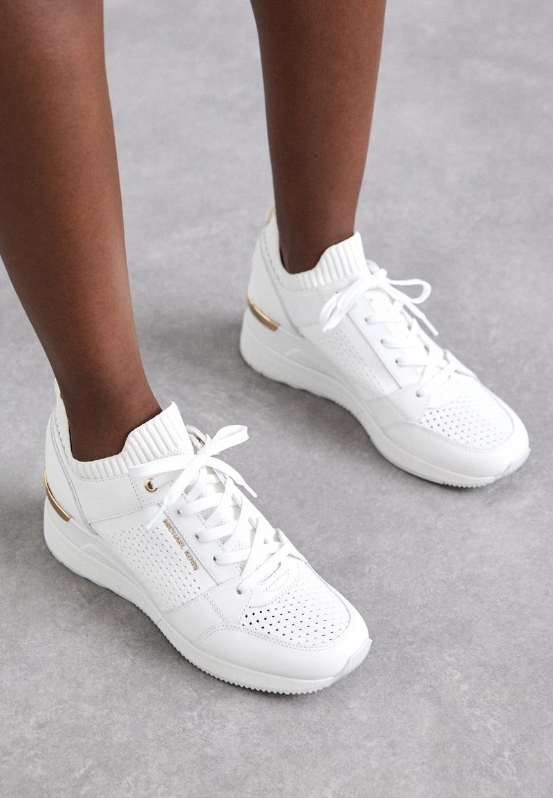 Personne portant des baskets blanches avec des détails perforés, des accents dorés et des lacets blancs sur un fond de sol gris.
