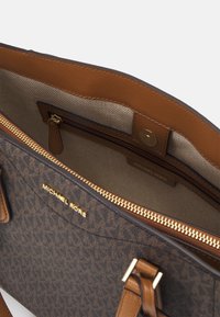 Sac fourre-tout brun avec logo imprimé, doté d'un zip sur le dessus, d'accents en cuir beige et d'une poche intérieure. Décoré d'un logo doré de la marque à l'intérieur.