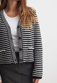 Cardigan noir et blanc rayé avec des manches longues, un col châle et des boutons en façade. Le tissu semble texturé avec une bordure contrastante.