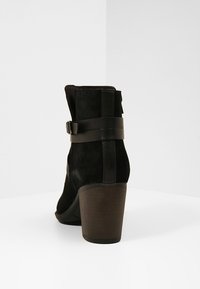 Musta mokkanahkainen nilkkuri paksulla korolla, jossa on sivuvarusteltu solkikiinnitys ja sileät nahkakoristeet, esitellen siistin, modernin muotoilun.