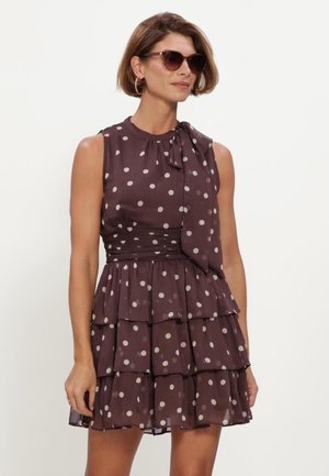 Vrouw draagt een mouwloze bruine jurk met witte stippen, een gelaagde rok en een strikdetail, gecombineerd met een zonnebril en creolen.