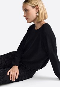 Black knit sweater with a round neck, paired with textured black pants adorned with floral patterns and sequins. Model seated at an angle.