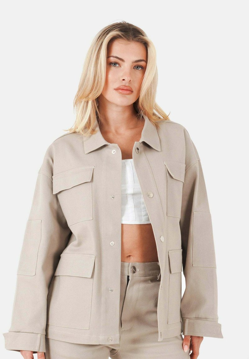 Blond kvinna i beige knappad jacka med framtida fickor över vit kortärmad topp och matchande beige byxor mot vit bakgrund.