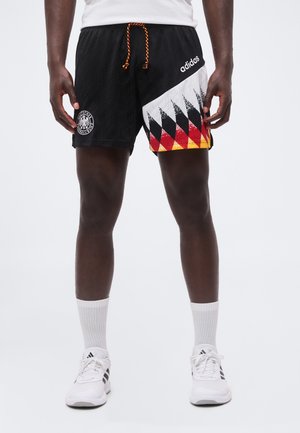 Άνδρας που φοράει μαύρα σορτς Adidas με μοτίβο της γερμανικής σημαίας και λευκά παπούτσια και κάλτσες Adidas, στέκεται μπροστά σε απλό φόντο.