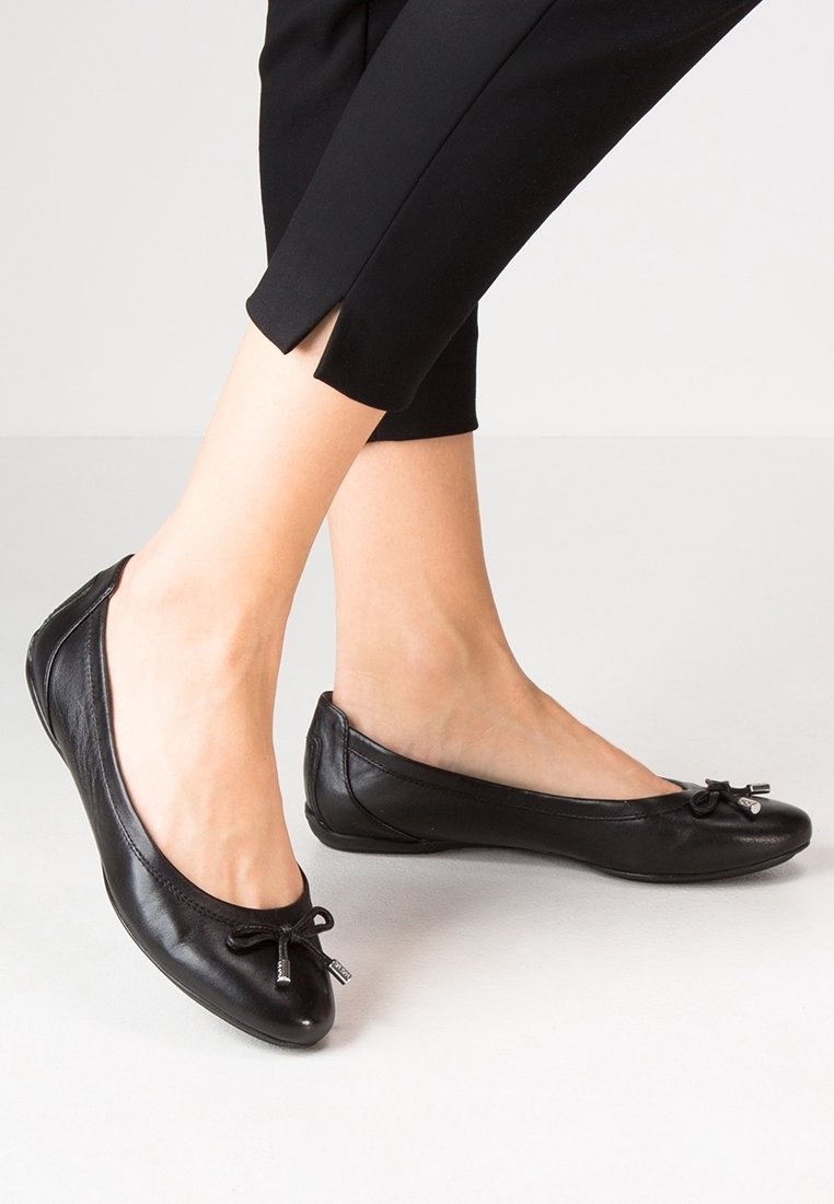 Femme portant des ballerines noires avec un petit détail nœud et un pantalon noir court, debout sur un sol clair.