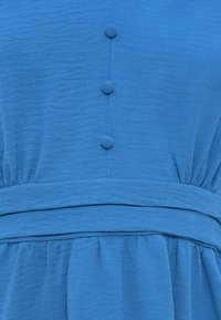Μπλε φόρεμα με υφή υφάσματος, που διαθέτει μια σειρά από τρία κουμπιά στο μπροστινό μέρος και ελαστική μέση με μαζεμένο ύφασμα από κάτω.