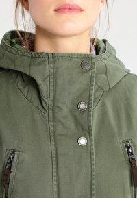 Zelená kapucí s mikinou se zapínáním na zip a patentními knoflíky, s prošívanou texturou a bočními kapsami na zip. Detaily tvoří stříbrné kovové prvky.