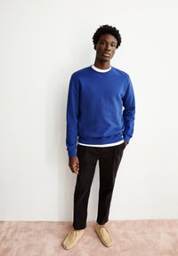 Blaues Sweatshirt mit gerippten Bündchen und Saum, über einem weißen Hemd getragen. Schwarze Hose und beigefarbene Slipper vervollständigen das Outfit. Einfarbiger Hintergrund.