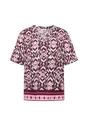 Blouse à manches courtes avec motifs géométriques bordeaux, blancs et roses, col à nouer avec pompons et bordure décorative à l'ourlet.