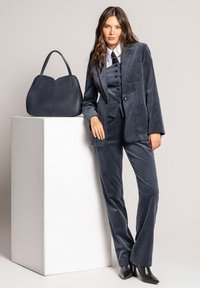 Kobieta w ciemnoniebieskim kostiumie z kordurowej tkaniny i czarnych butach opiera się na białym postumencie, na którym leży duża ciemnoniebieska torebka na tle jednolitego tła.