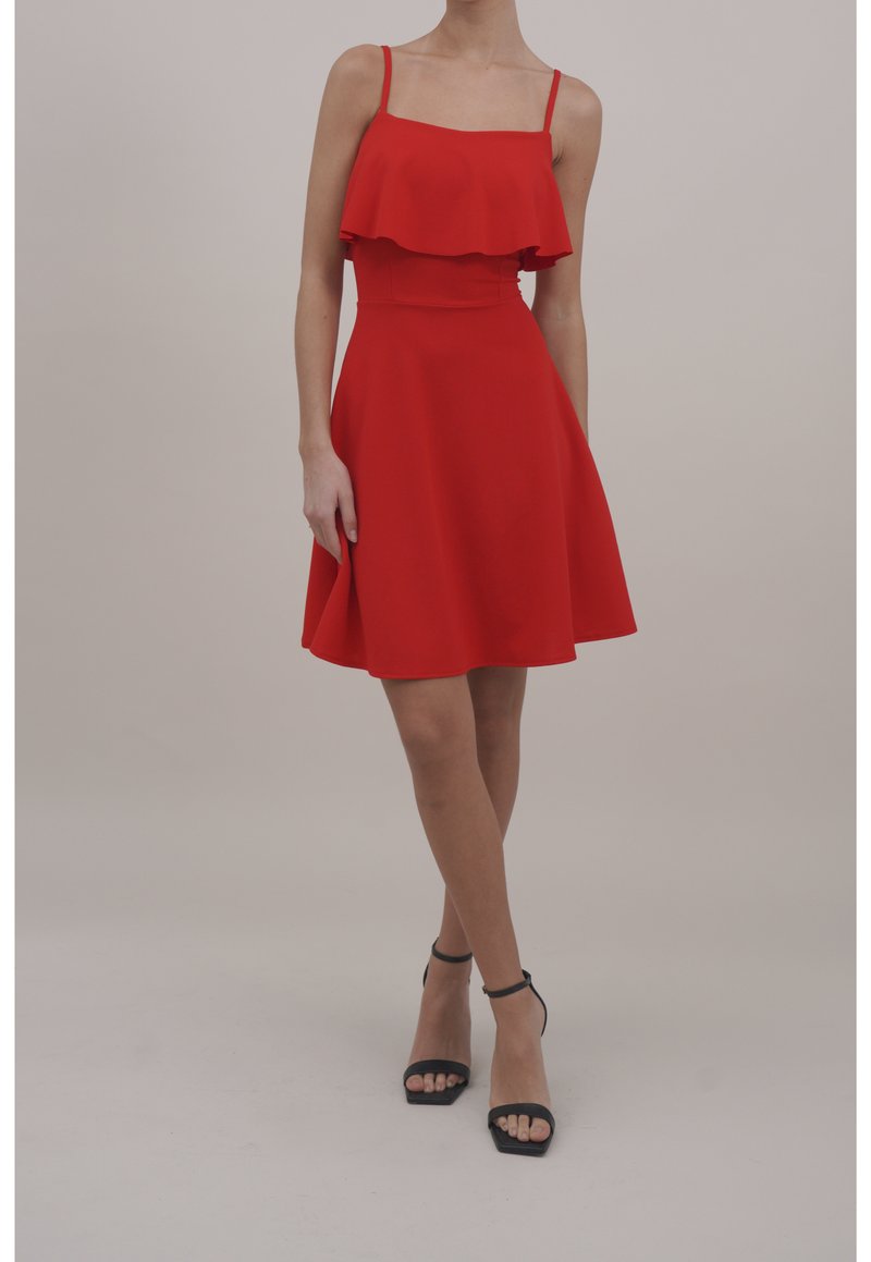 Femme portant une robe rouge sans manches avec un corsage à volants et des sandales noires à talons hauts à bride cheville, debout devant un fond uni.