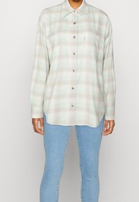 Camicia a quadri chiara in tessuto morbido, con colori verde e beige, colletto a punta e chiusura con bottoni sul davanti, dotata di due tasche sul petto.