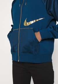 Person som bär en blå zip-up hoodie med guld Nike-logotyp och fickor, händer i fickorna, i kombination med svarta byxor.