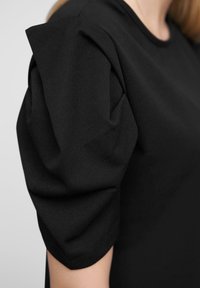 Haut noir à manches courtes présentant des manches structurées et bouffantes. Le tissu est lisse avec un léger brillant, et le col est arrondi.