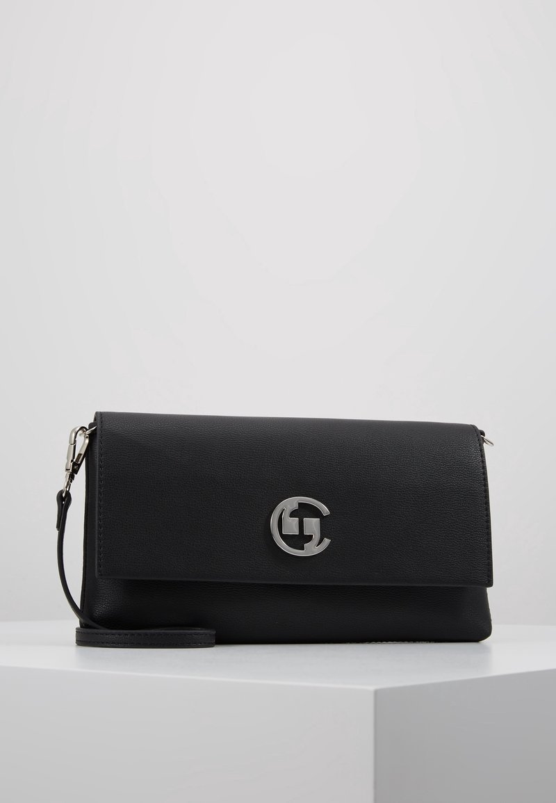 Pochette en cuir noir avec logo circulaire argenté et bandoulière détachable, posée sur une surface blanche sur un fond clair uni.