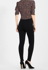 Mujer vestida con una blusa de manga abullonada de flores, jeans skinny negros y zapatos de tacón negro, de pie dando la espalda sobre un fondo blanco.