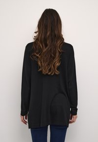 Maglione nero con maniche lunghe, vestibilità ampia e polsini a costine. Texture liscia e bordo posteriore più lungo. Indossato con jeans blu scuro.