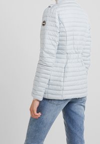 Světle modrá péřová bunda s prošívanou texturou, přiléhavým střihem a nastavitelným pasem. Obsahuje kulatou nášivku s logem na levém rameni.